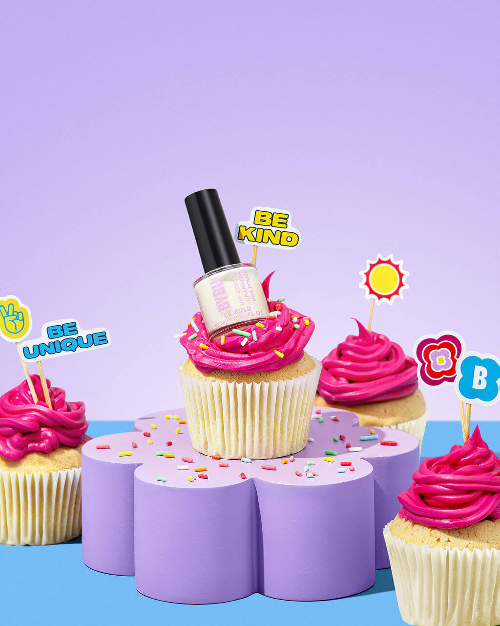Cupcakes with pink frosting and a bottle of nail polish on a purple stand against a light purple background.